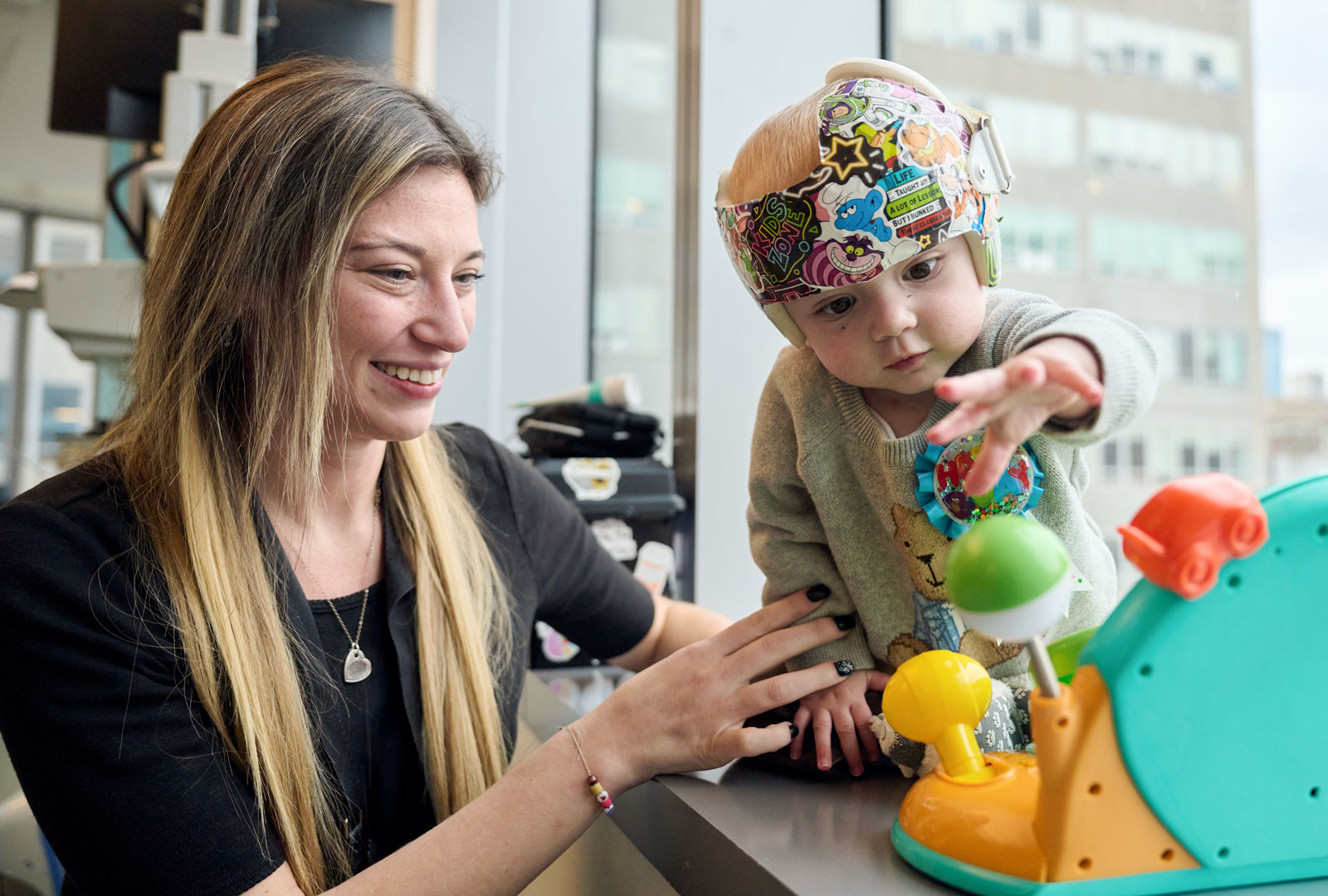 Smiling provider lightly holding and watching over child wearing helmet and playing with toy