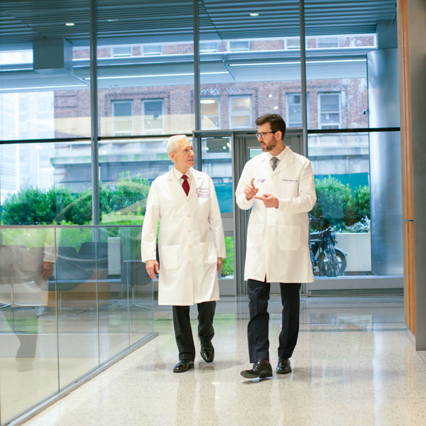 Two doctors in white coats talking, with one gesturing with his hands, while walking inside of a glass-front building
