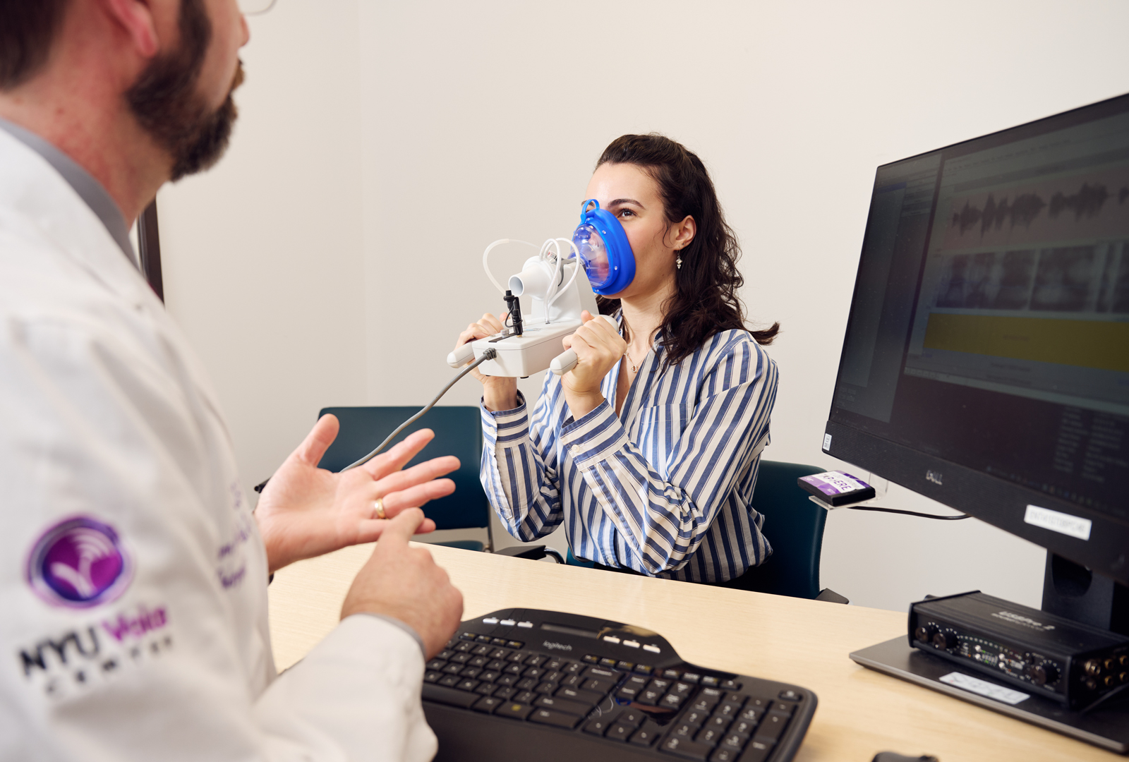 Provider looks on at patient wearing an apparatus around their mouth that is sending readouts about their voice to a computer